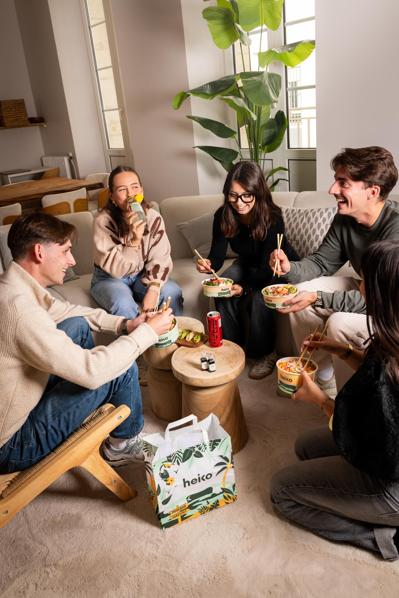 Cinq amis assis ensemble dans un salon moderne, mangeant des plats de poke avec des baguettes et buvant des boissons.
