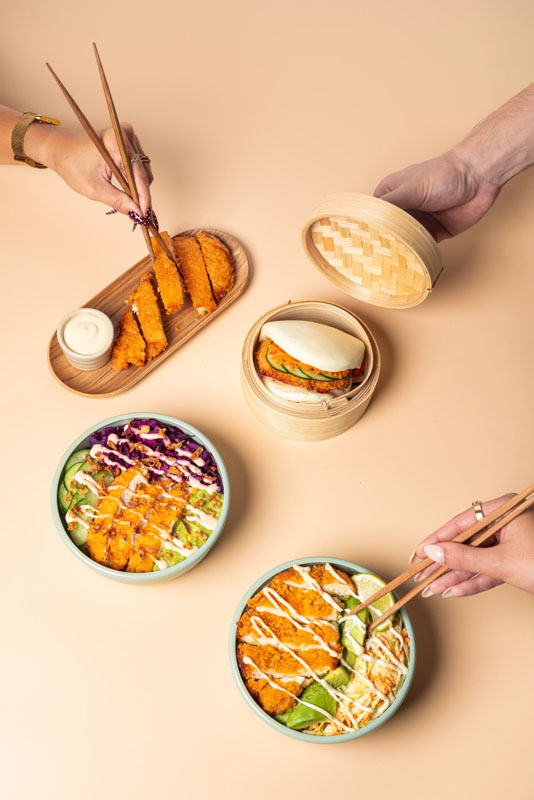 Deux bols de poke bowl avec du poulet pané, des concombres, de la laitue et une sauce mayo, un pain bao avec un morceau de poulet, et des mains utilisant des baguettes pour prendre des morceaux de poulet pané accompagnés de sauce mayo.