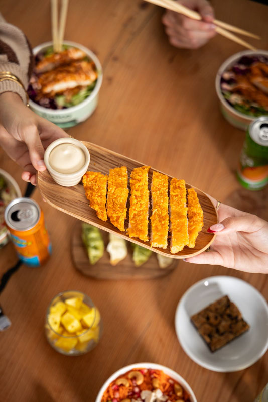 Deux mains tenant un plateau en bois avec des tranches de poulet pané croustillant et une petite coupelle de sauce blanche, sur une table avec divers plats et boissons.
