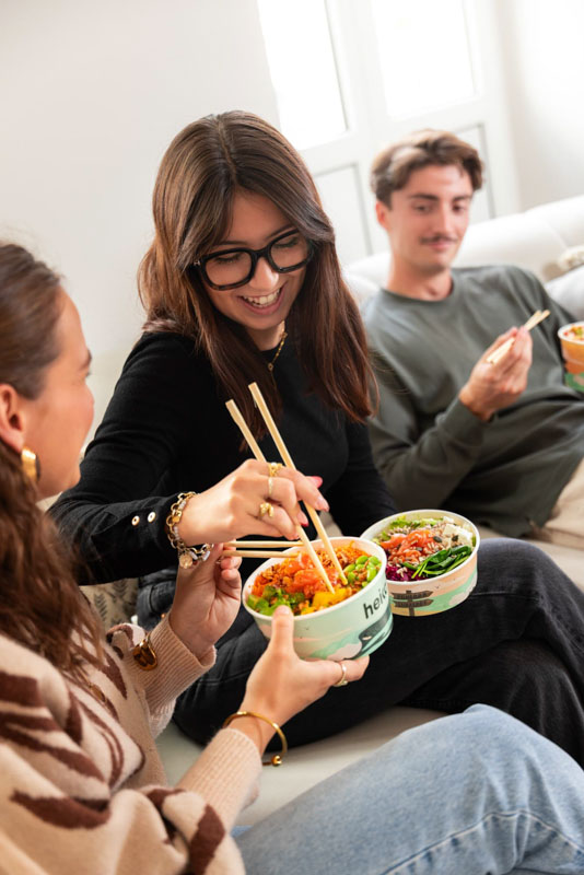 Trois jeunes adultes assis sur un canapé, mangeant des bols poke avec des baguettes et souriant.