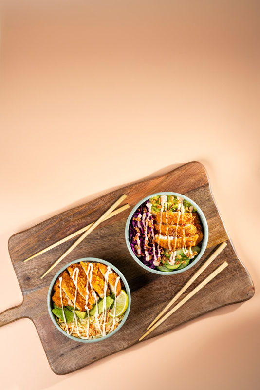 Deux bols de poke garnis de filet de poulet grillé, légumes frais et sauce, servis sur une planche en bois avec deux paires de baguettes.