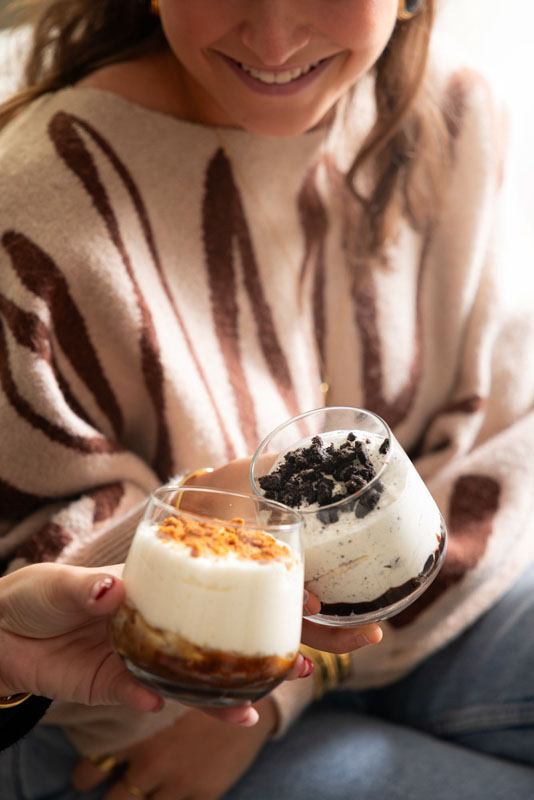Femme souriante tenant un verre de dessert avec des morceaux de biscuits, trinquant avec une autre personne tenant un verre de dessert à la crème et crumble.