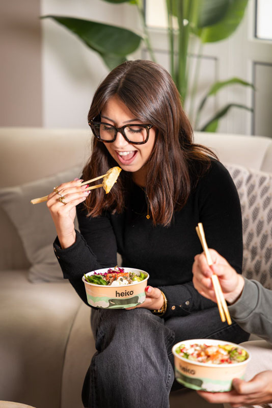 Jeune femme souriante mangeant avec des baguettes un bol de nourriture saine marqué 'heiko' dans un salon moderne.