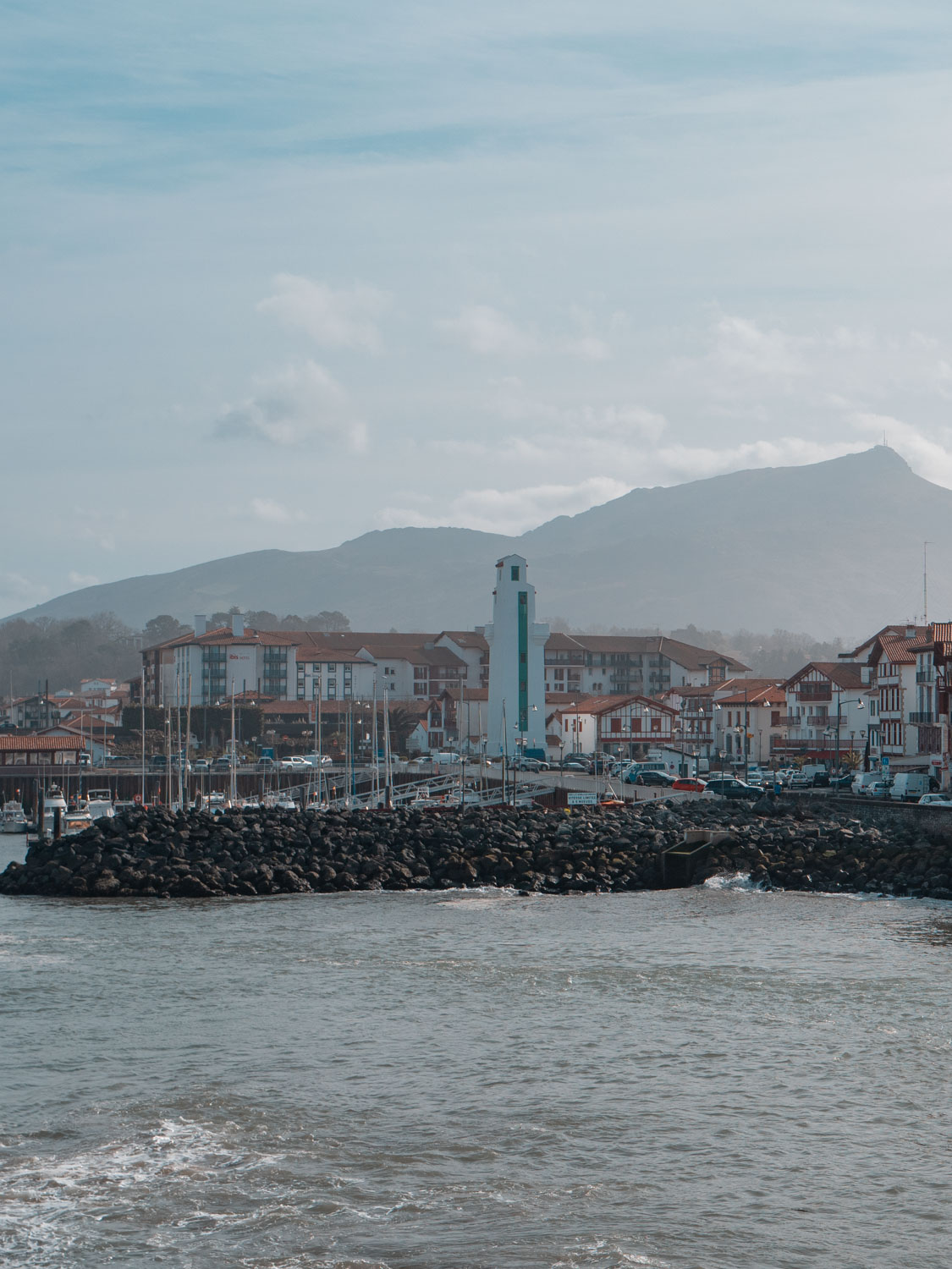 Vue du port avec des bateaux amarrés, des bâtiments à toits rouges et une montagne en arrière-plan sous un ciel légèrement nuageux.