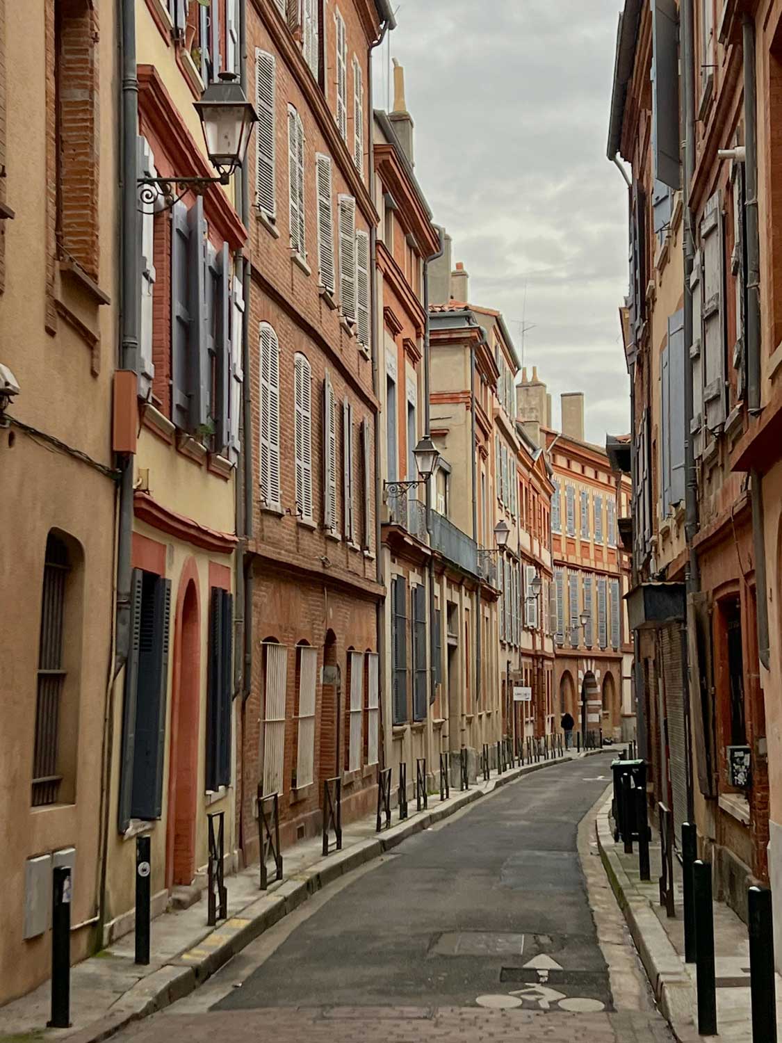 Rue étroite bordée de bâtiments en briques rouges avec volets, surplombée par un ciel nuageux.