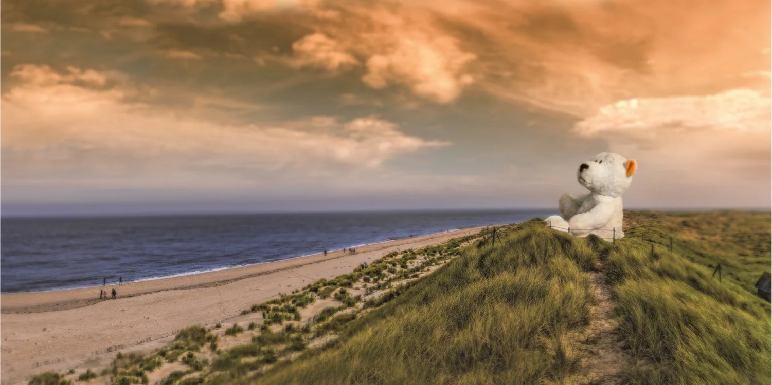 A building sized teddy bears sits on the top of a sand dune and watches over the sea with the sun setting in the back behind a group of clouds.