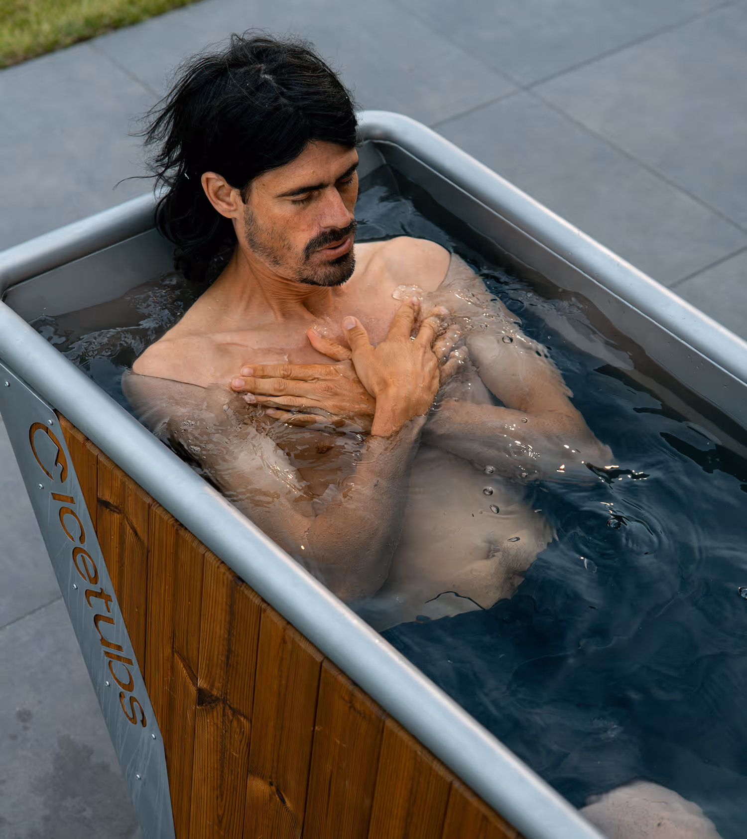 Man with dark hair and beard sits in a wooden ice bath tub outdoors with hands crossed on chest.