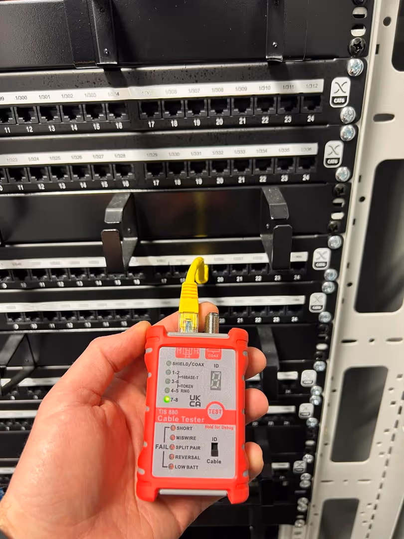 A hand holding a red cable tester with a connected yellow wire, in front of a server rack.