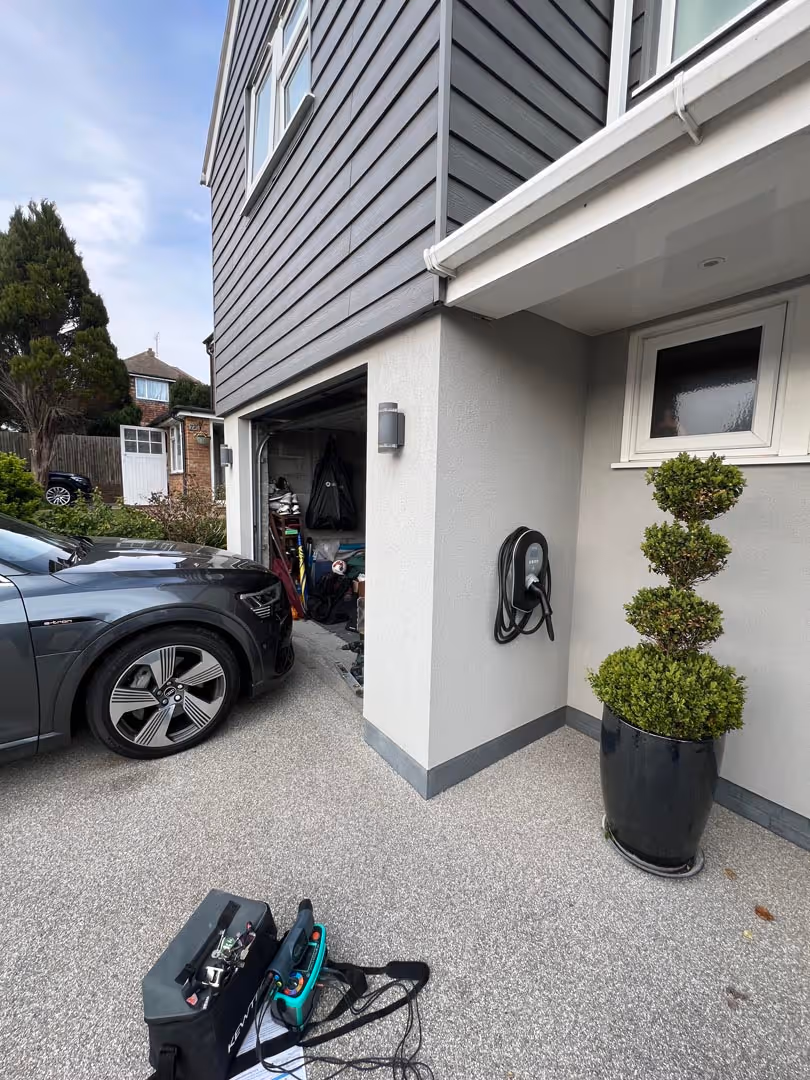 Car parked next to a house with charging equipment and a planter by the garage.