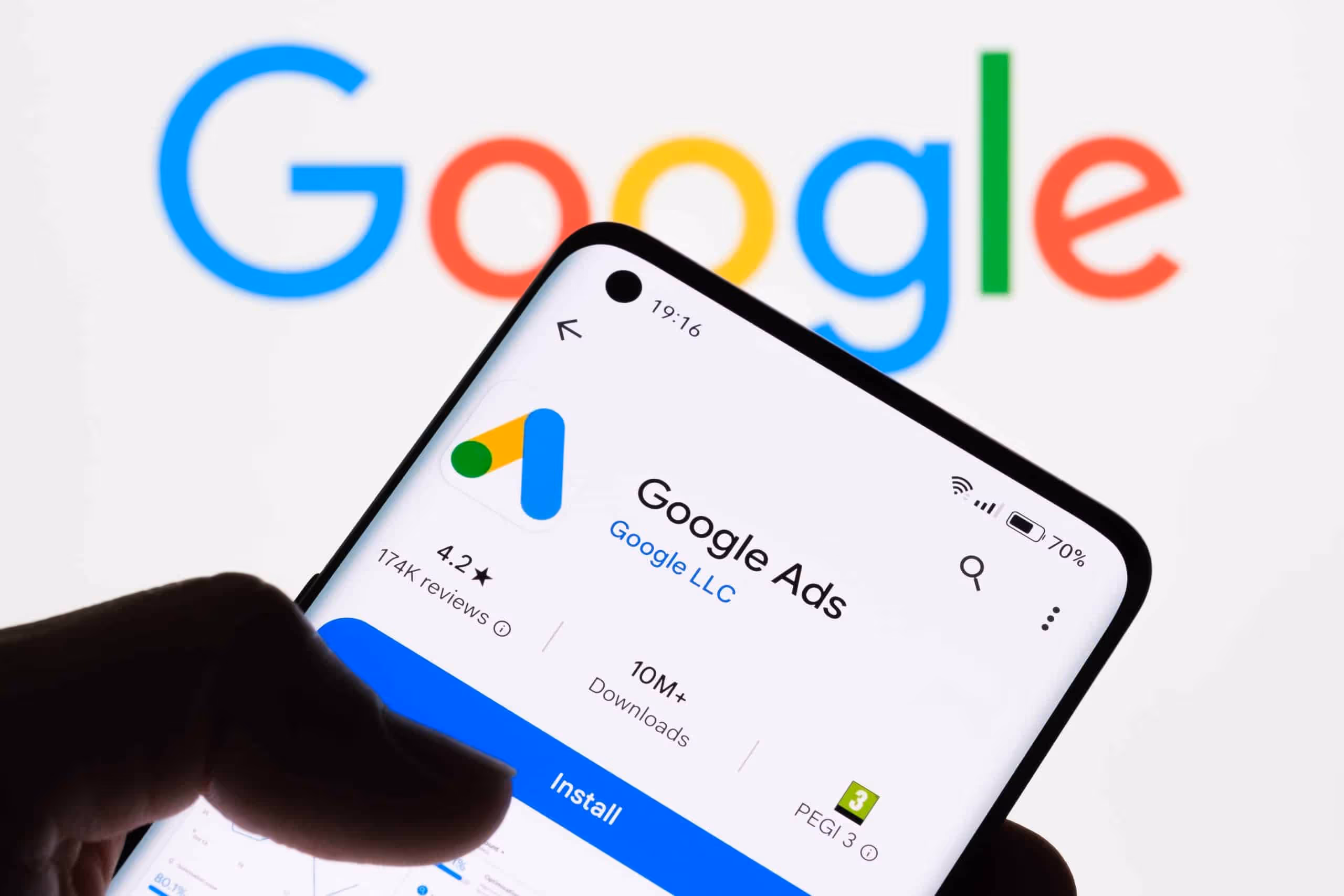 Hand holding a smartphone displaying the Google Ads app page, with the Google logo in the background.