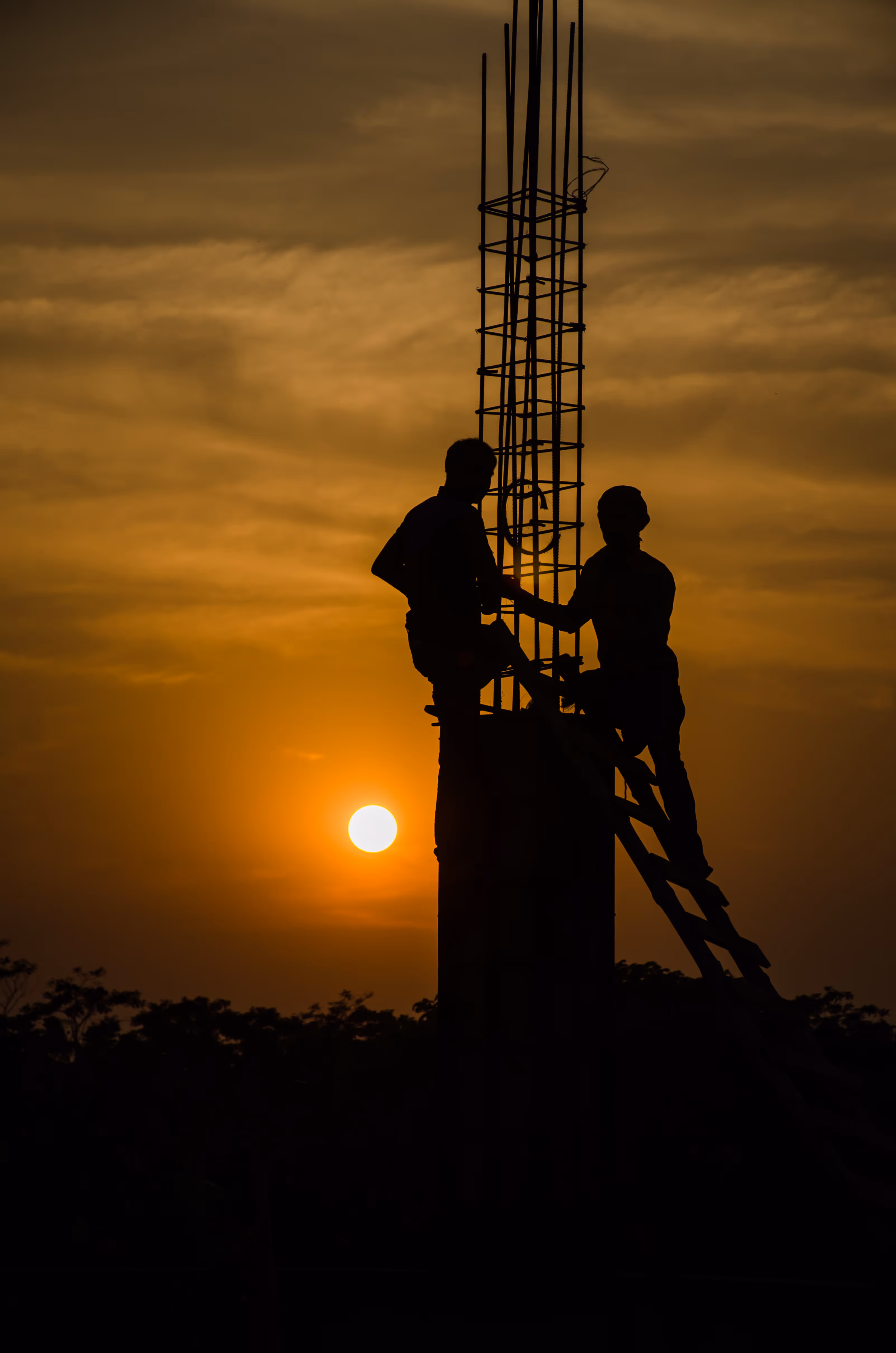 A silhouetted image of two construction workers standing on a structure with metal rods, against an orange sky with the sun setting behind them.