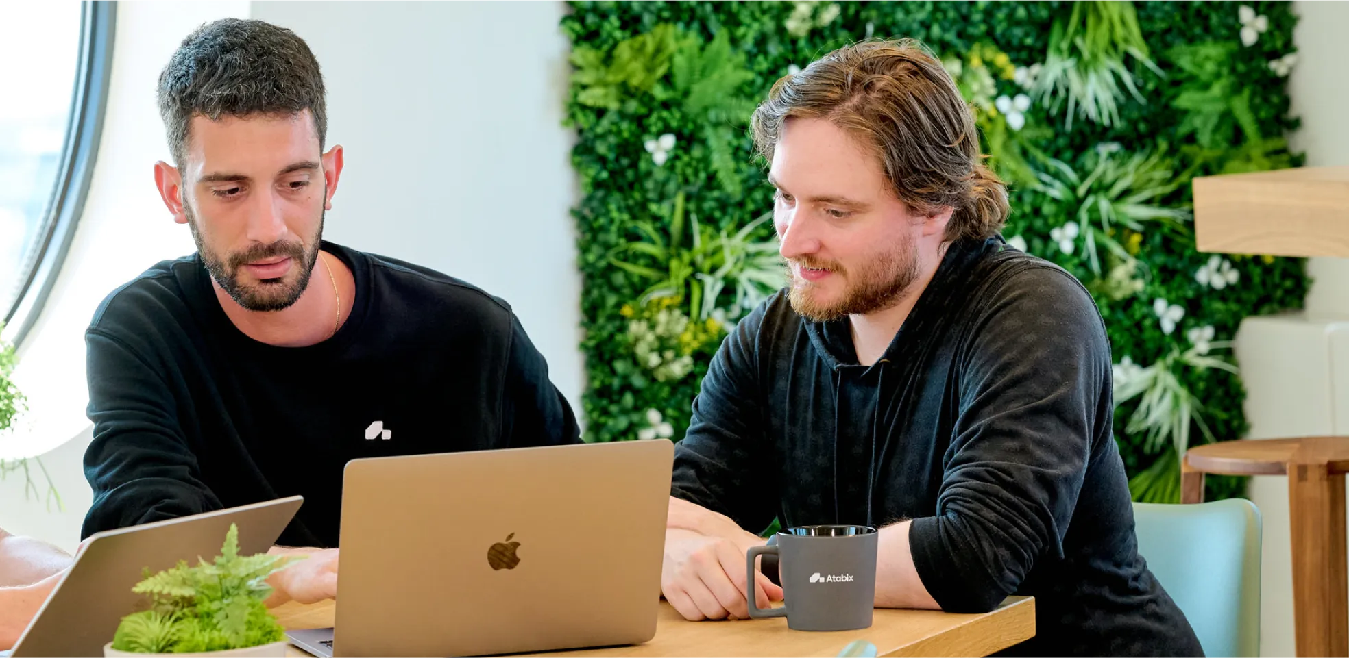 Twee mannen die naar een laptop kijken in een kantoorruimte met veel planten.