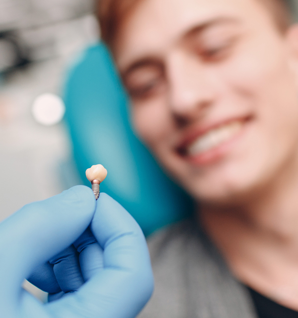 Gloved hand holding a small dental implant with a smiling patient blurred in the background.