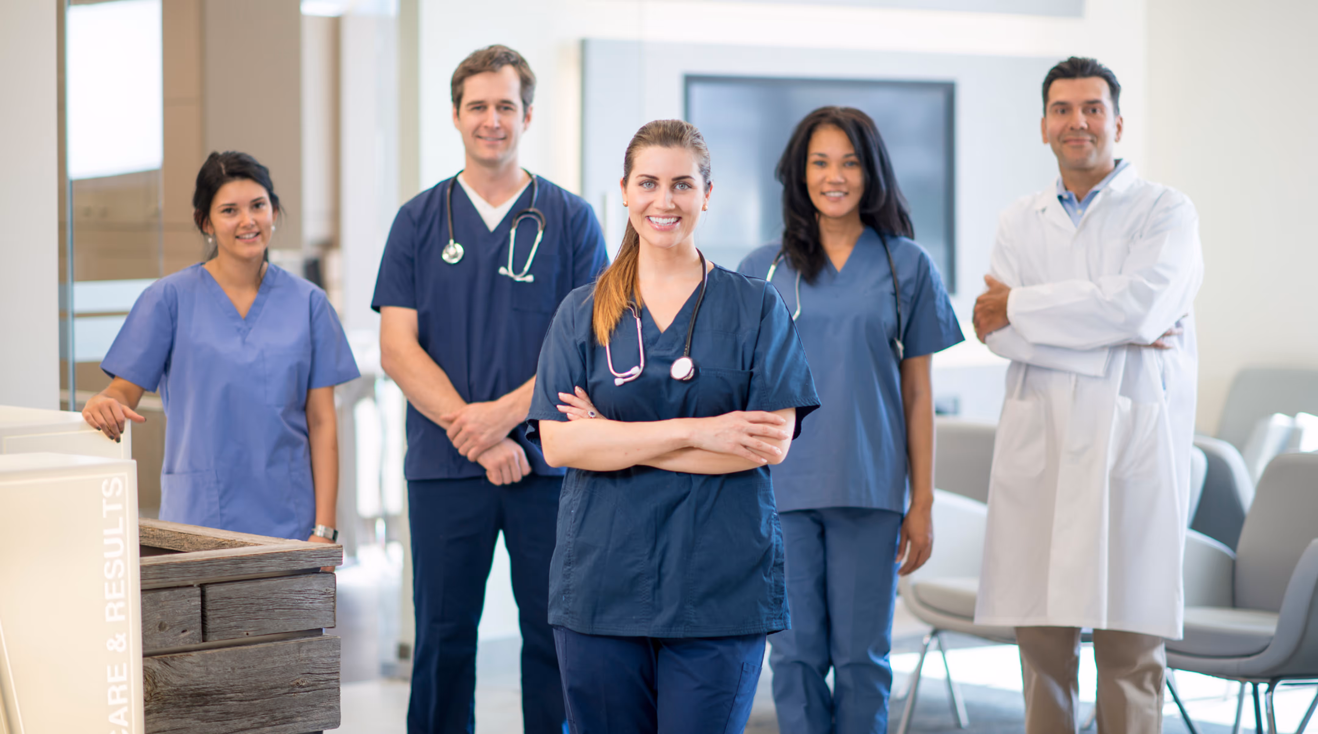 Group of five diverse medical professionals in scrubs and a white coat smiling in a modern medical facility.