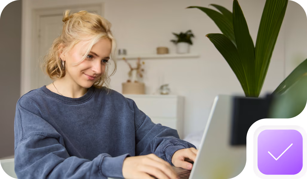 A woman with blonde hair tied back, wearing a blue sweatshirt, sitting at a table and working on a laptop in a bright room with a large green plant nearby.