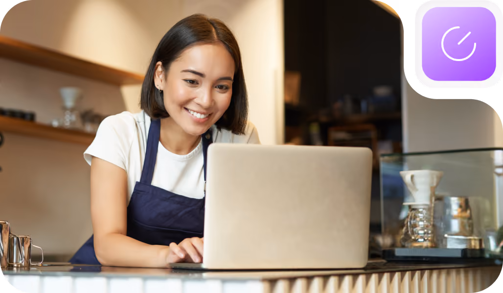 Smiling woman wearing a dark apron leans on a counter while working on a laptop in a cozy cafe setting with shelves and coffee equipment in the background.