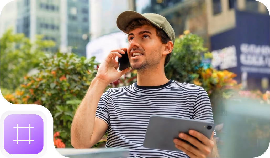 A man wearing a striped shirt and green cap is sitting outdoors, holding a tablet in one hand and talking on a smartphone, with blurred urban buildings and greenery in the background.