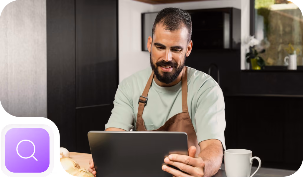 A bearded man wearing a brown apron and light green shirt is sitting at a kitchen table, smiling and looking at a tablet in his hands, with a white mug next to him.