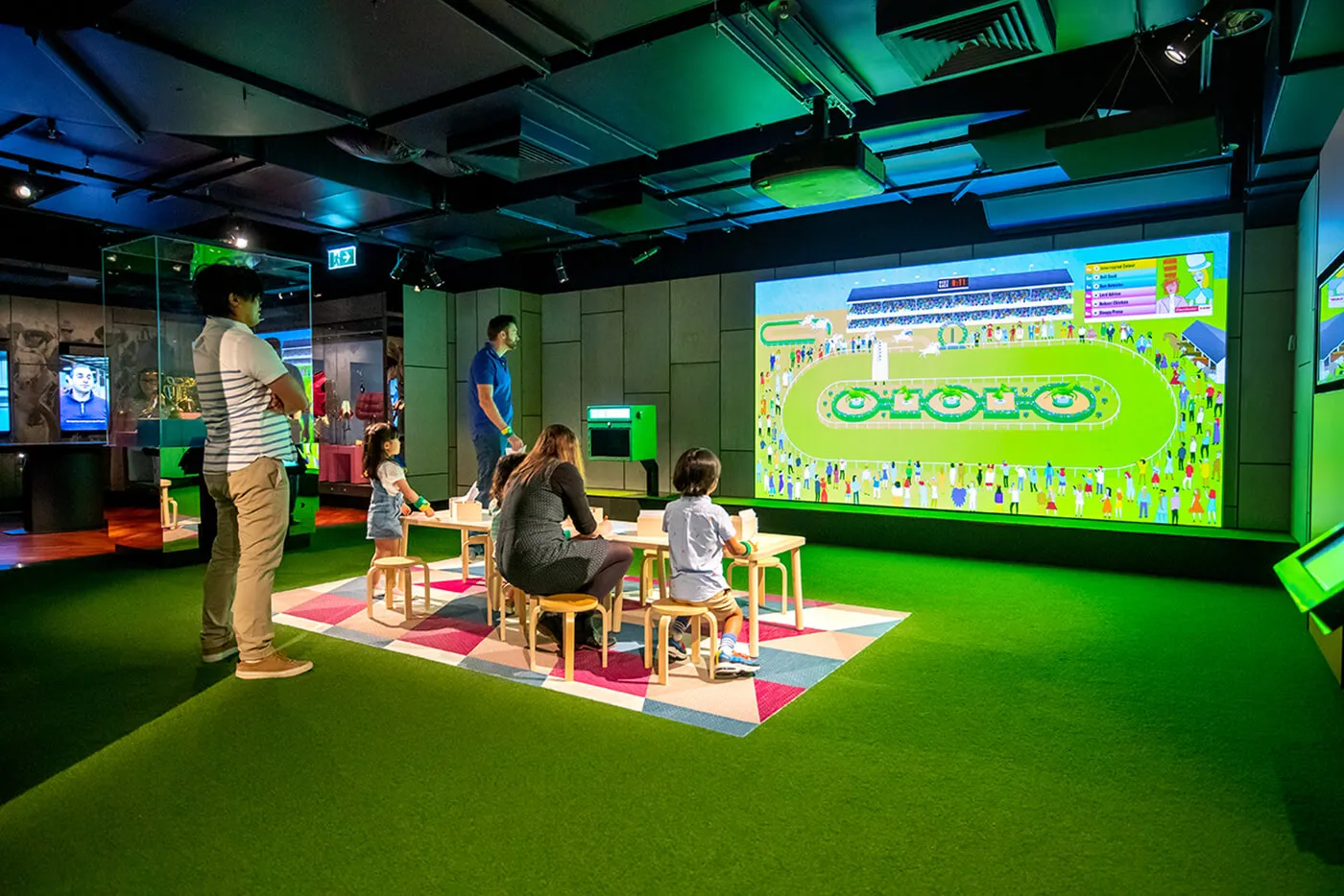 Group of people are standing around colouring tables, or colouring in template. Race occurring on large screen.