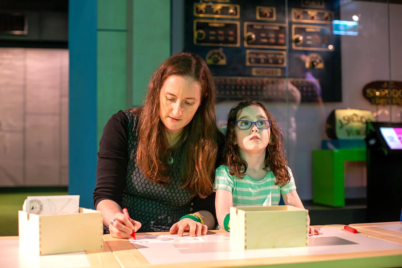 A woman colouring in a horse template, and her daughter beside her stares at the screen where a race is taking place.