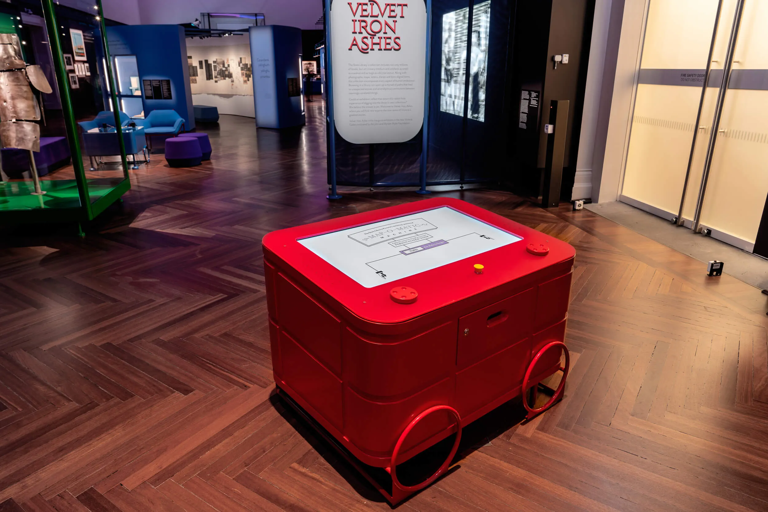 Bright red Map-O-Matic machine in the gallery. The idle screen says "The State Library Victoria's amazing, patented Map-O-Matic machine. Explore the connections through your very own personalised tour. Rotate the dials to begin."