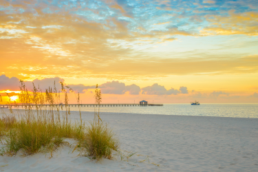 Sea oats grow on sandy dunes beside a calm beach at sunset, with a long pier and a small boat in the distance.