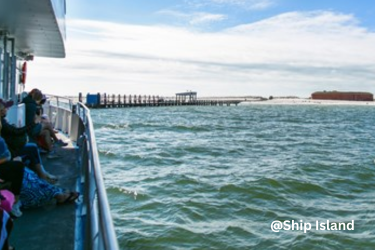 Passengers sit along a ferry’s open deck as it approaches Ship Island, with a pier and brick fort visible across the water.