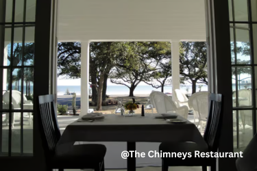 A table set for two at The Chimneys Restaurant overlooks a porch, oak trees, and the waterfront through open French doors.