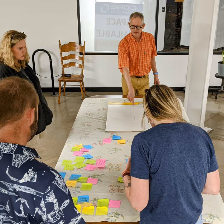 Instructor leading a workshop with participants gathered around a table covered with papers and colorful sticky notes.