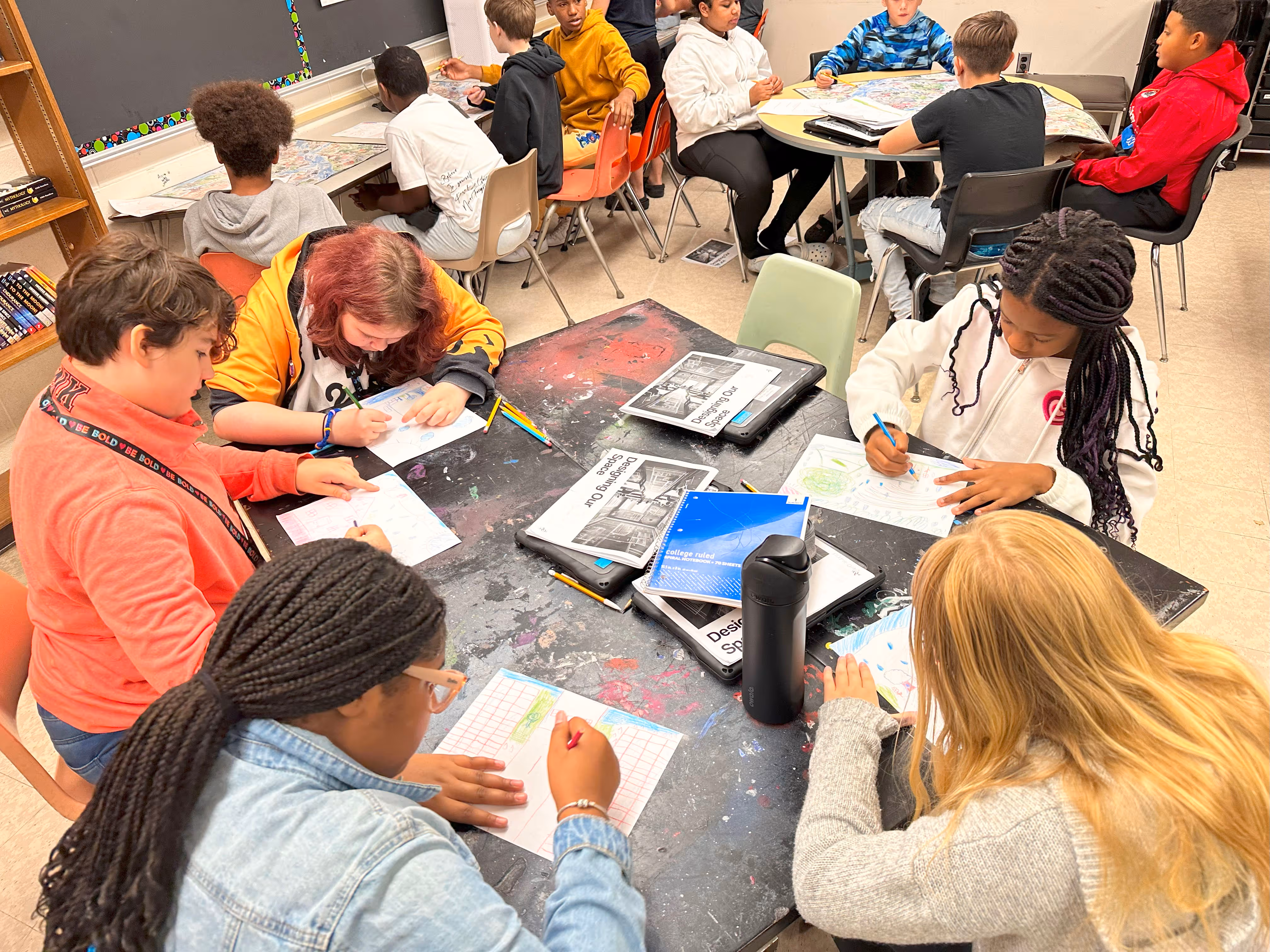 Students working collaboratively at a table with papers, tablets, and materials in a classroom setting.