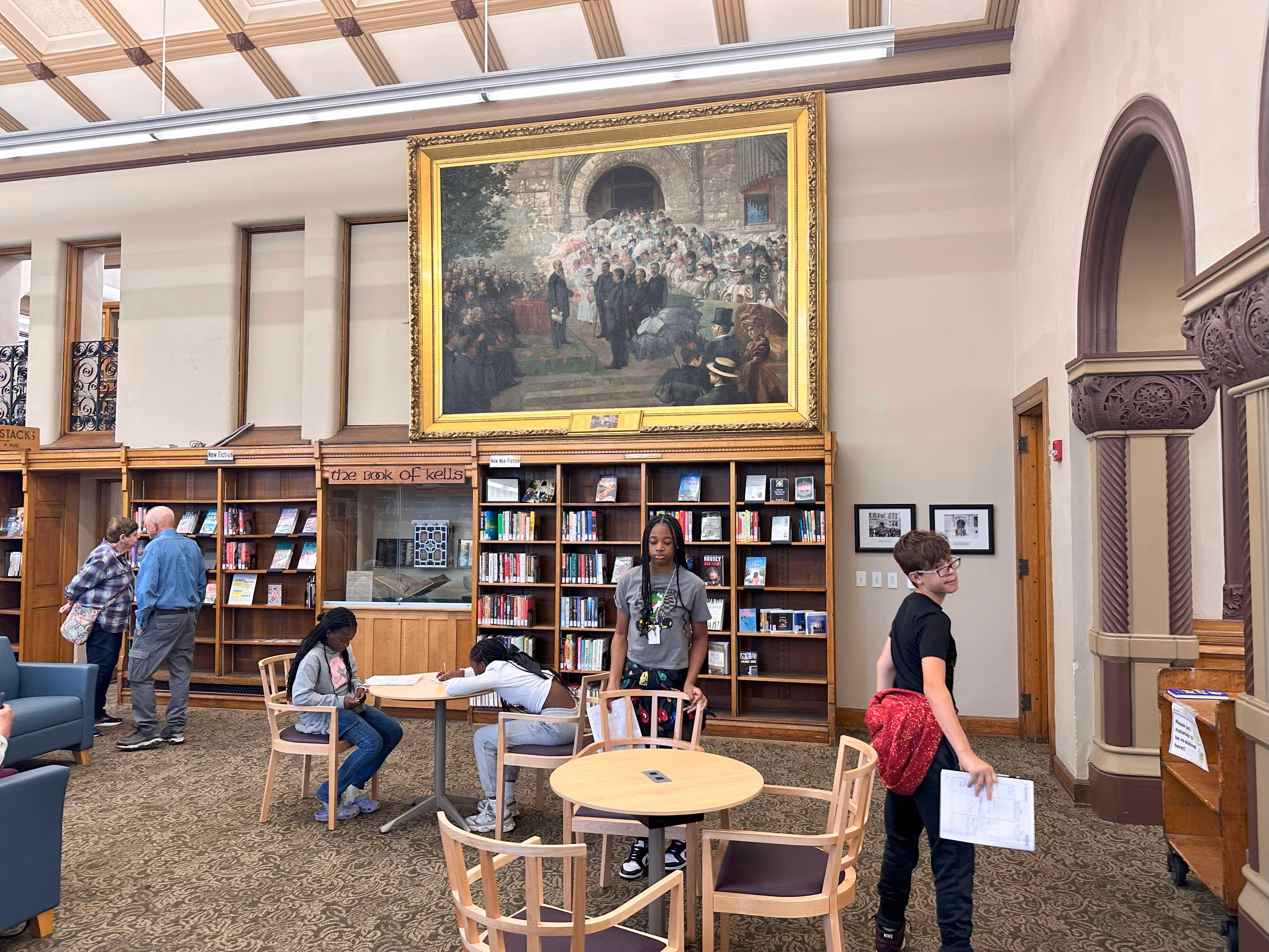 People studying and working in a library with tall bookshelves and a large historical painting on the wall.