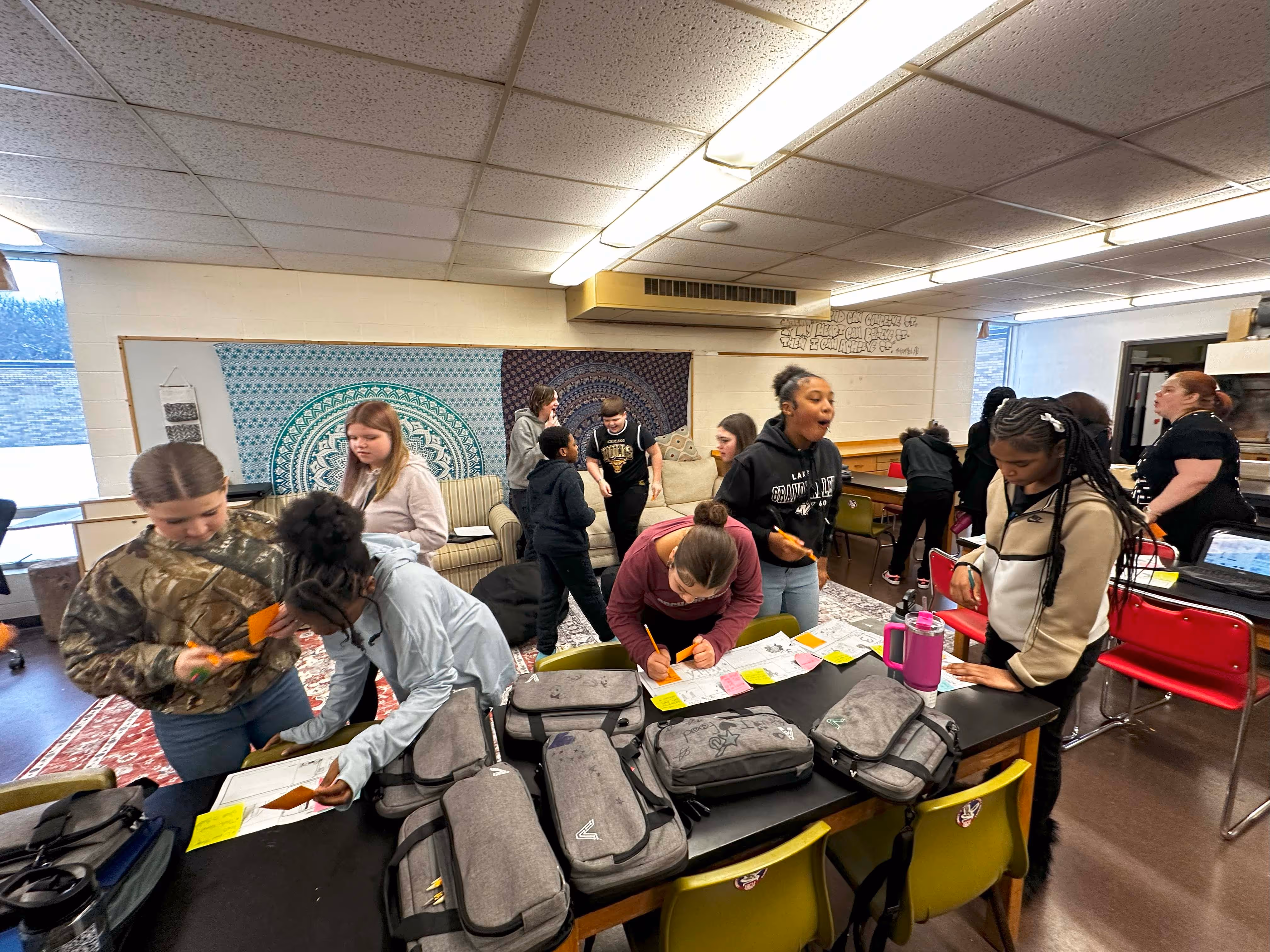 Students gathered around tables working on projects with papers and supplies in a classroom.