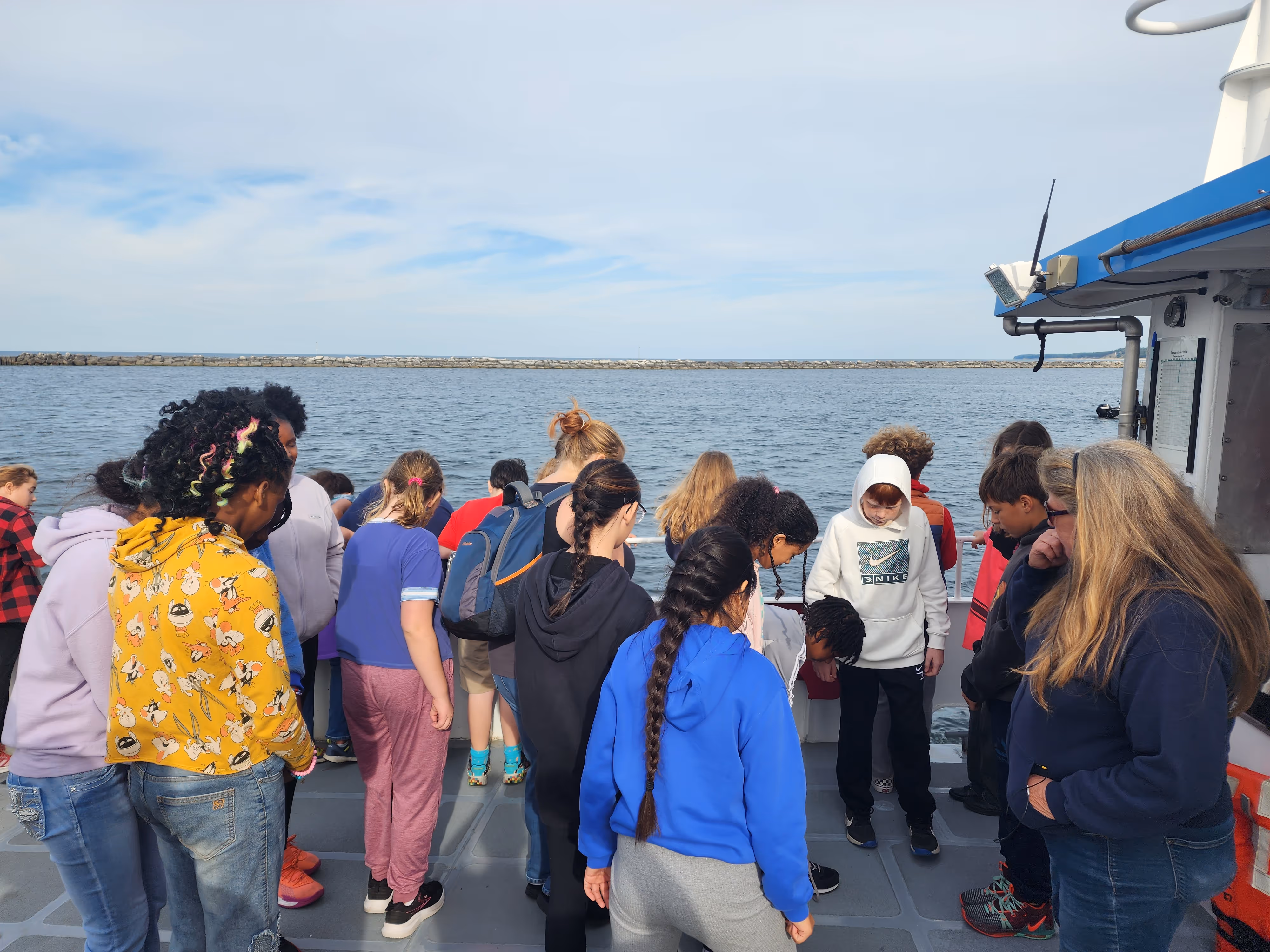 Group of diverse children and an adult standing on a boat deck looking down towards the water under a partly cloudy sky.