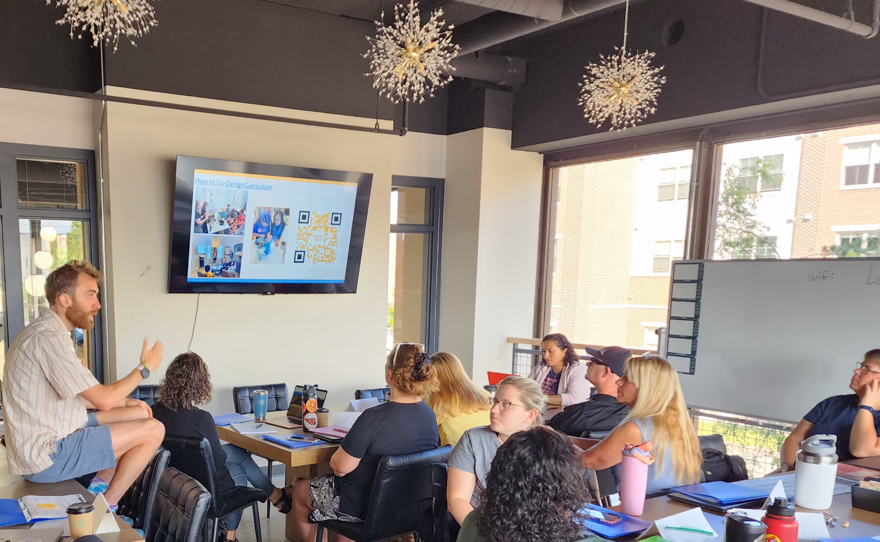 A man presenting to a group seated around a conference table with a screen displaying 'How to Co-Design Curriculum' with photos and a QR code.