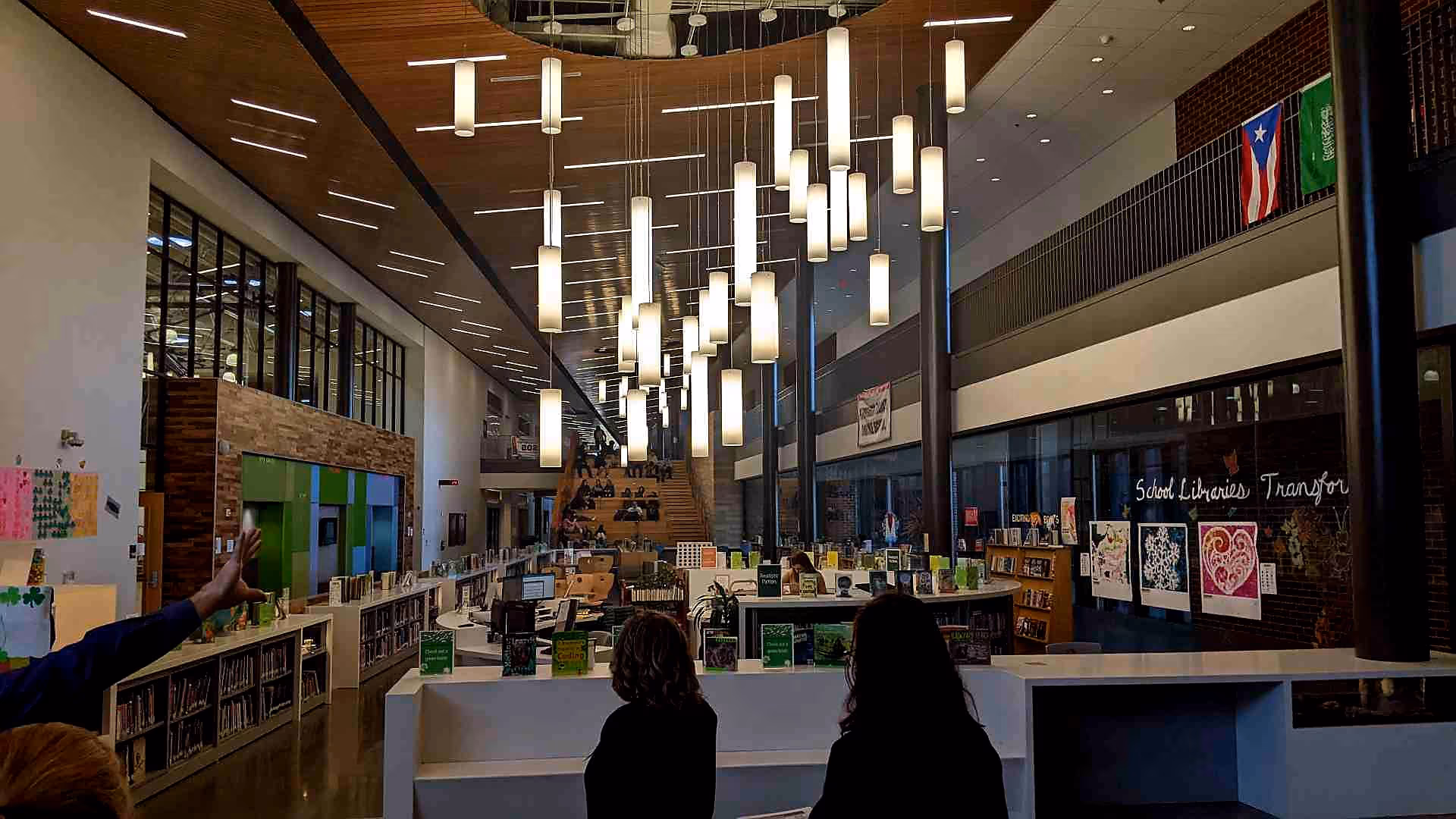 Inside a modern library with high ceilings, hanging cylindrical lights, bookshelves, artwork on glass walls, and people seated on stairs in the background.