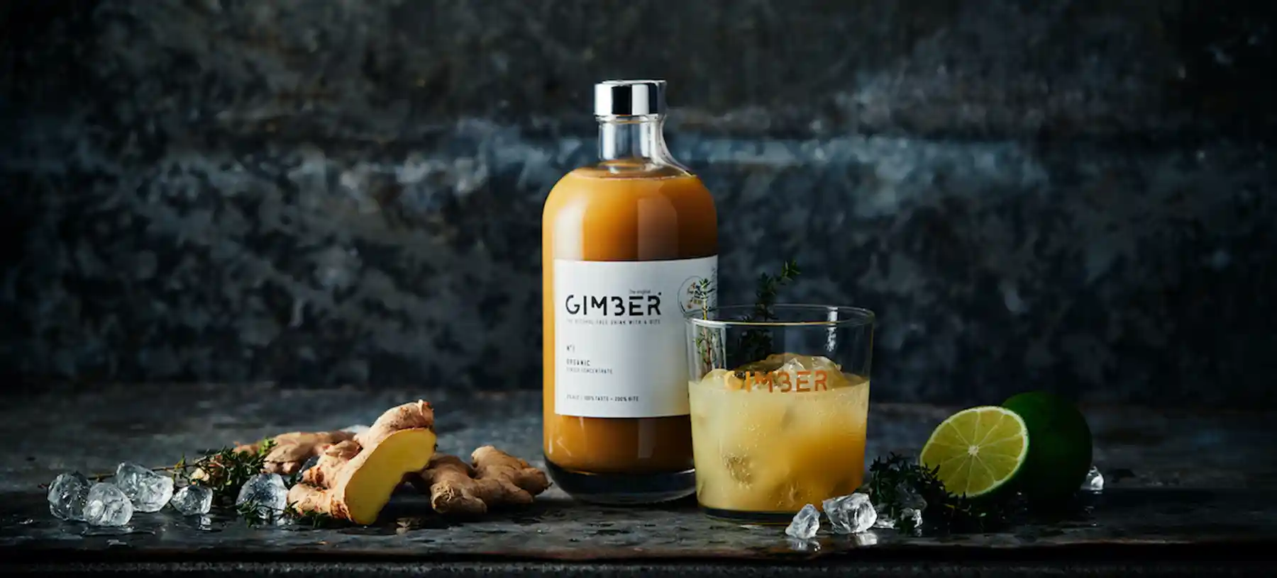 Ingwerdrink von GIMBER im Glas und in der gebrandeten Flasche vor dunkler Wand