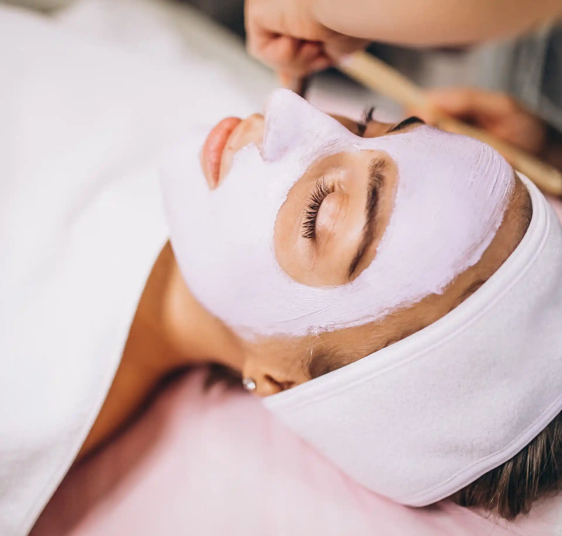 Frau im Spa mit Creme-Maske im Gesicht und mit Handtuch zugedeckt