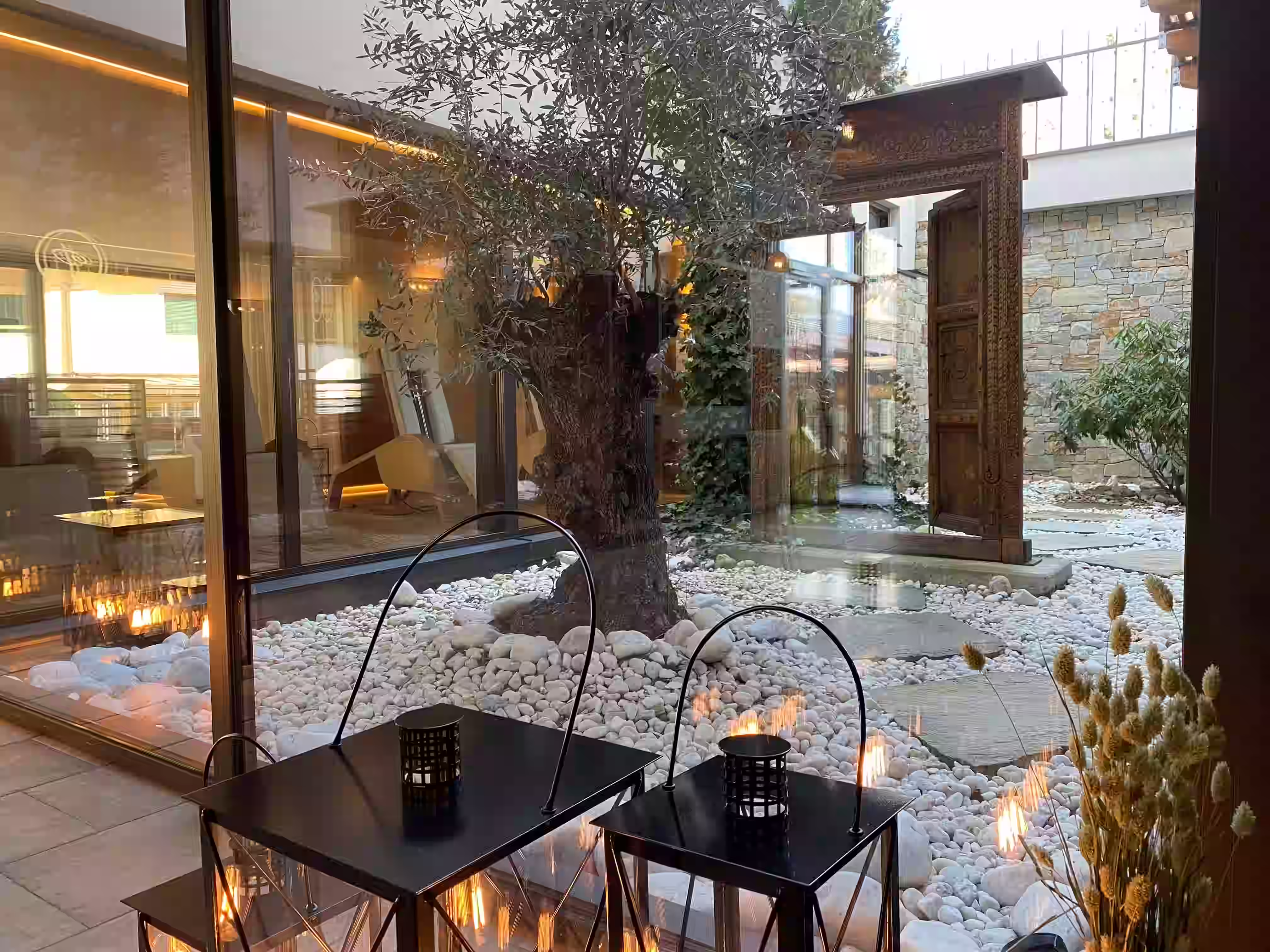 DAS PARADIES – Spa-Innenhof mit Olivenbaum, weißen Kieselsteinen, Laternen und antiker Holztür; Lounge im Hintergrund
