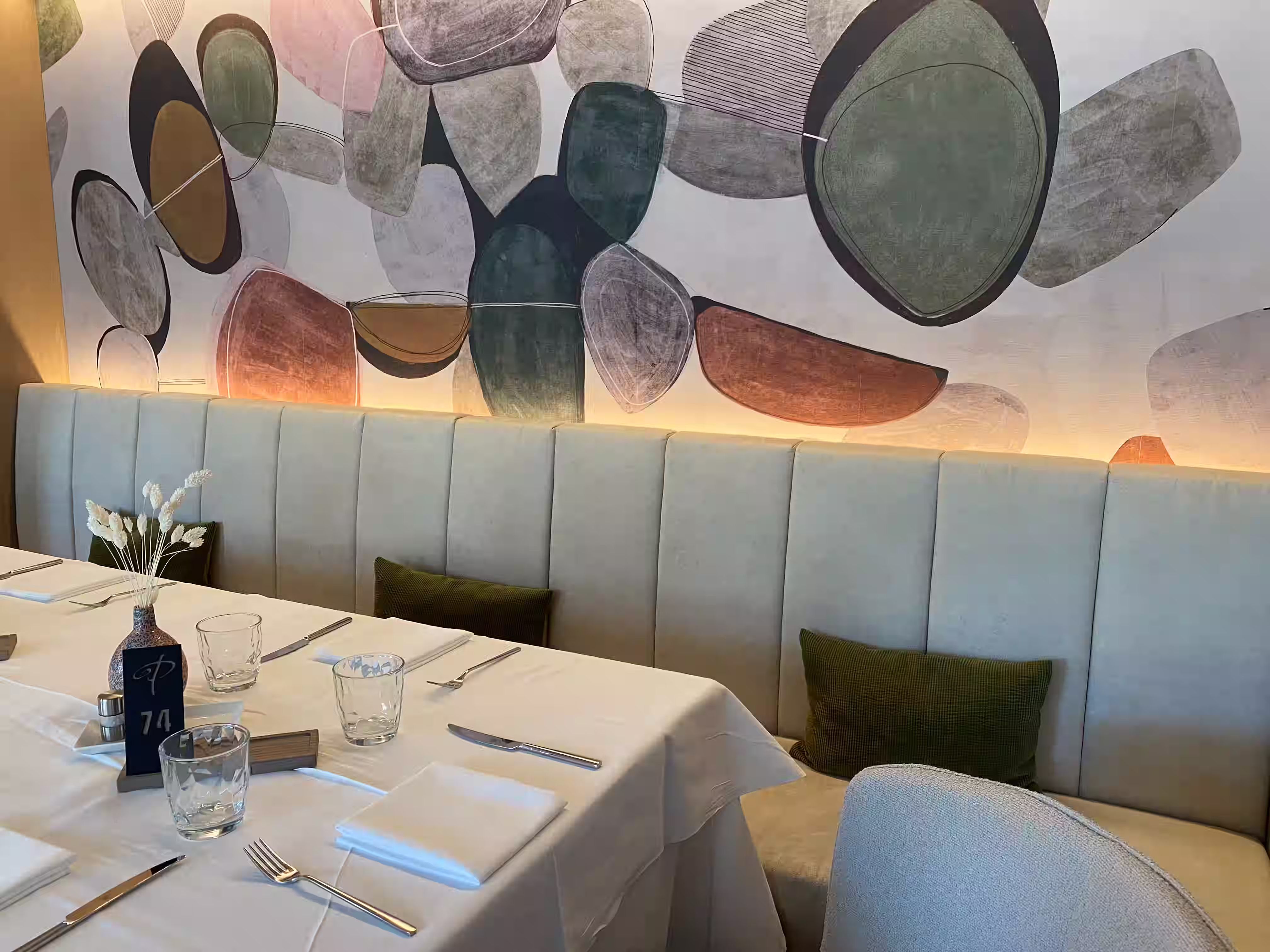 DAS PARADIES – Gedeckter Restauranttisch mit weißer Tischdecke vor einem großformatigen abstrakten Wandbild mit organischen Formen