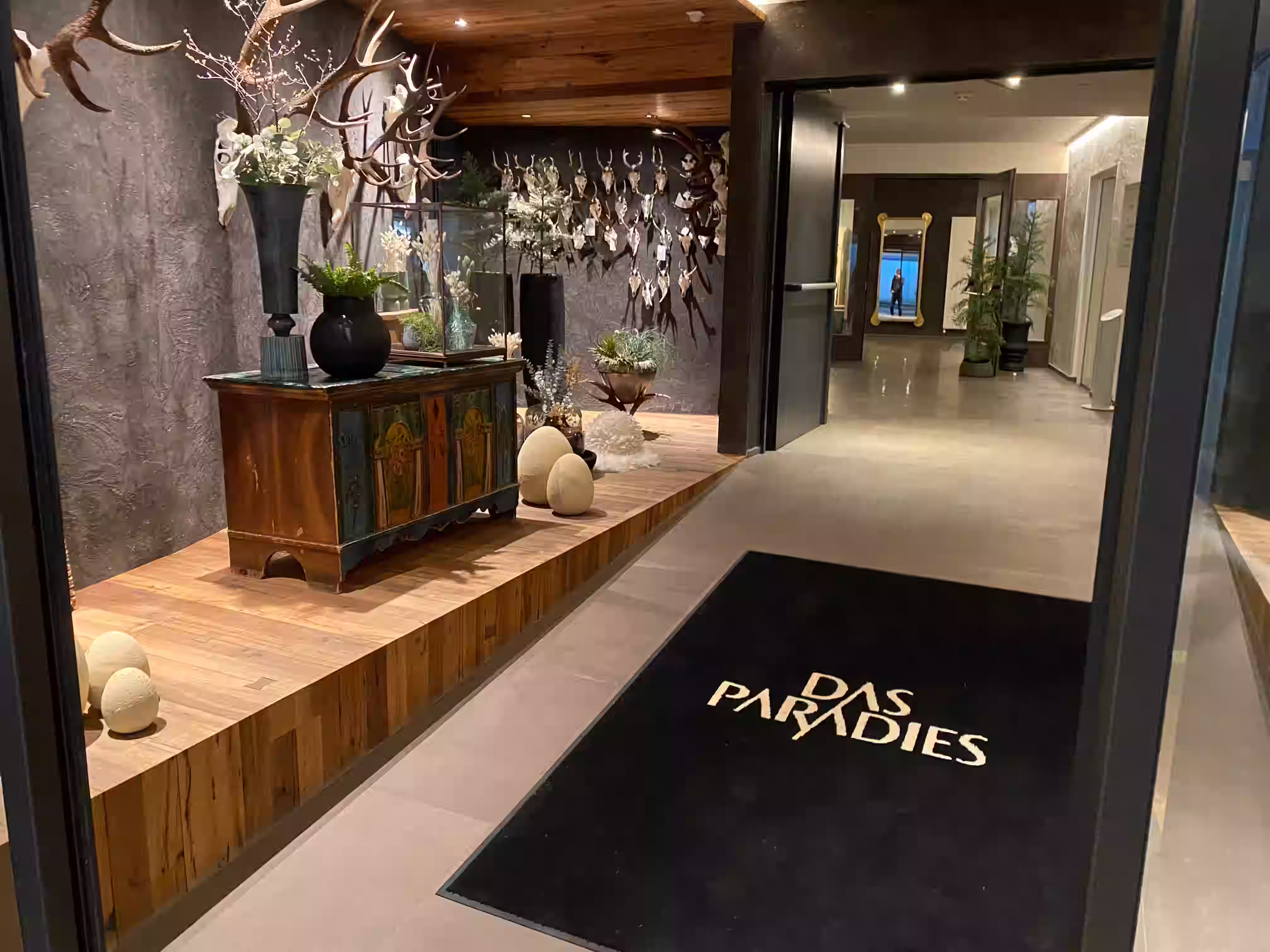 DAS PARADIES – Hoteleingang vom Parkhaus mit Logo-Fußabtreter, bemaltem Bauernschrank, Geweihinstallation und Blick in den Hotelkorridor