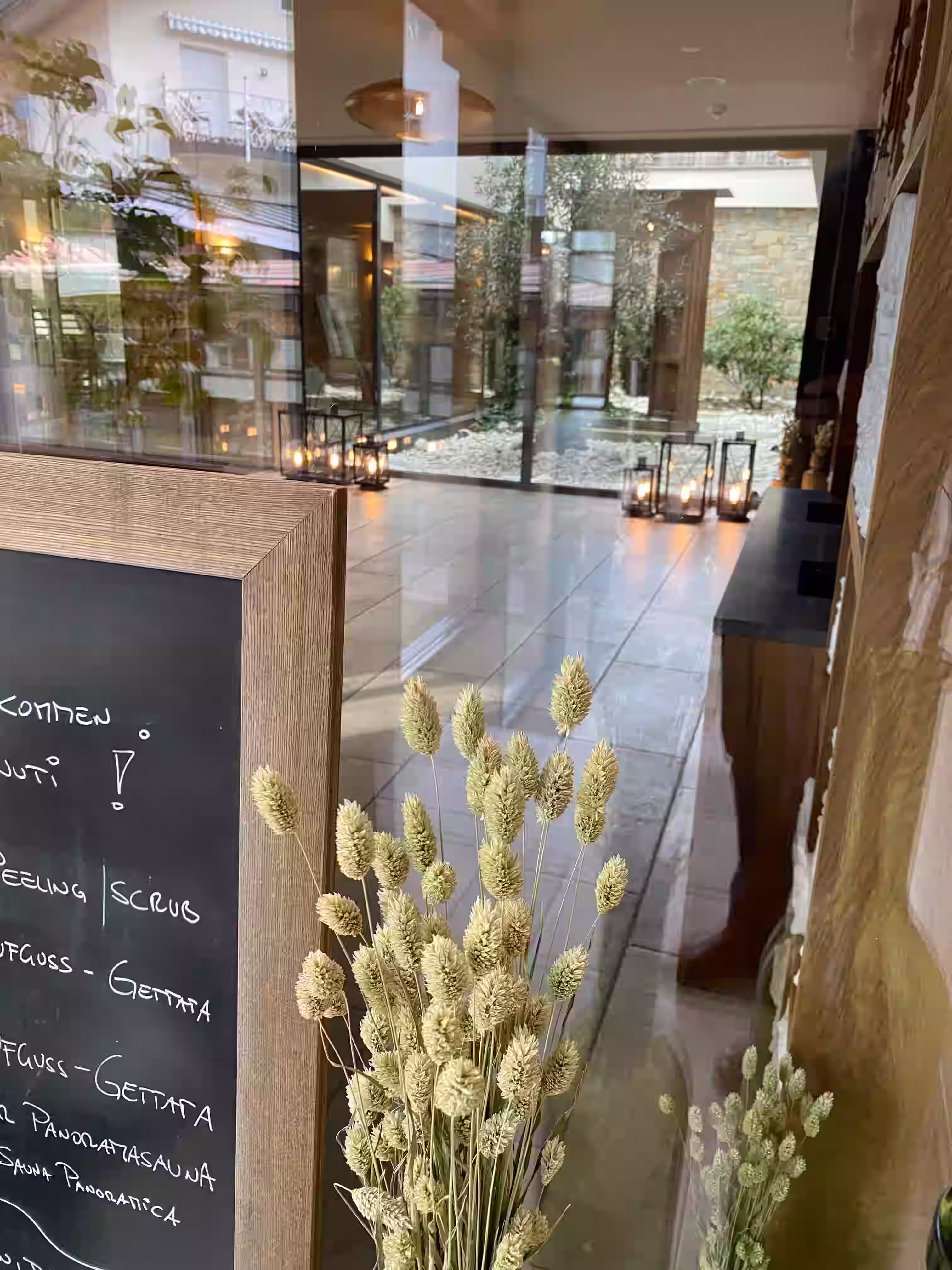 DAS PARADIES – Spa-Eingang mit handgeschriebener Kreidetafel und Tagesprogramm, Trockenblumen im Vordergrund, Blick auf den beleuchteten Innenhof mit Olivenbaum