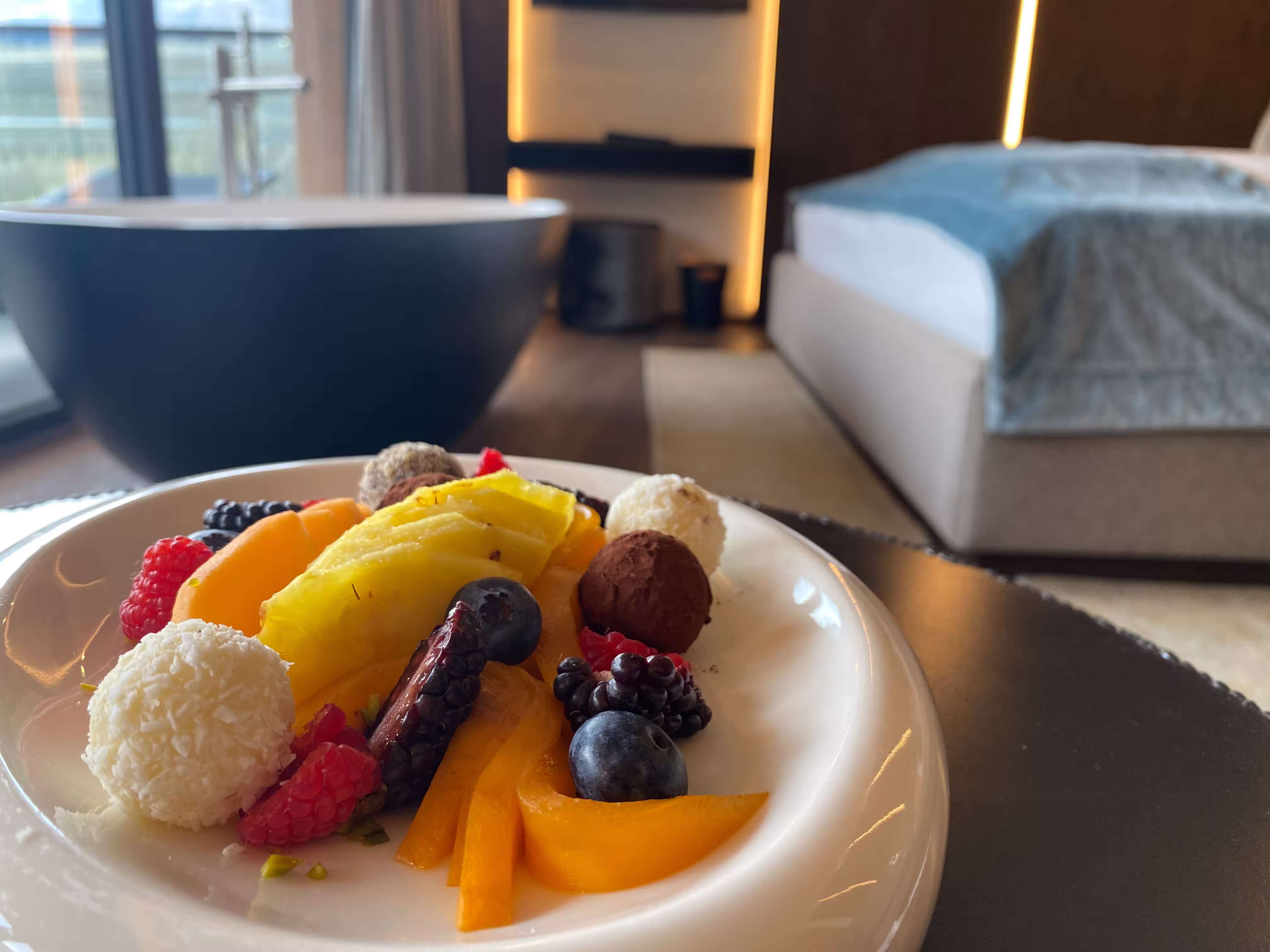 DAS PARADIES – Begrüßungsteller mit frischen Früchten und Pralinen im Zimmer; im Hintergrund freistehende Badewanne und Doppelbett