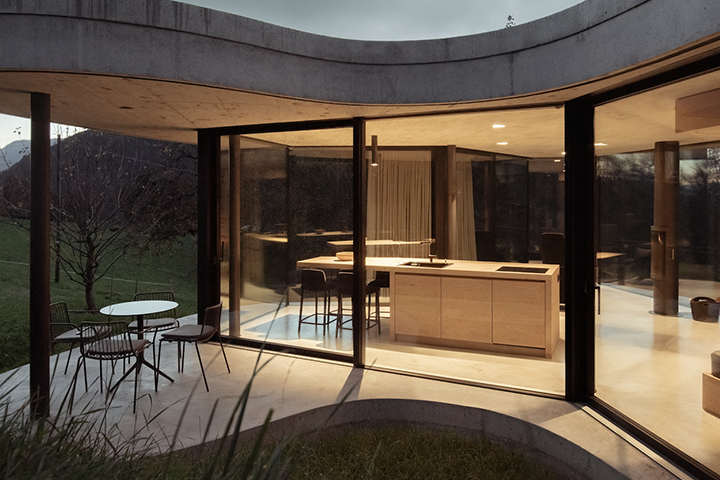 Freiform Guestouse in Südtirol. Sicht auf das Gebäude mit gesschwungener Architektur und durchgängiger Fensterfront in der Abenddämmerung, im Guesthouse Blick auf erleuchtete Küche