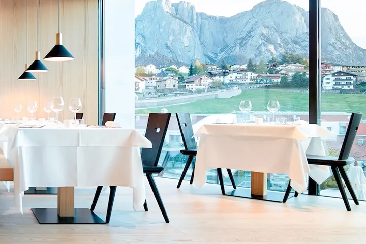 Hotel Schgaguler in Kastelruth. Restaurant mit TIschdecken und schwarzen Stühlen vor heller Holzwand und großer Fensterfront mit Blick auf Dorf und Berge