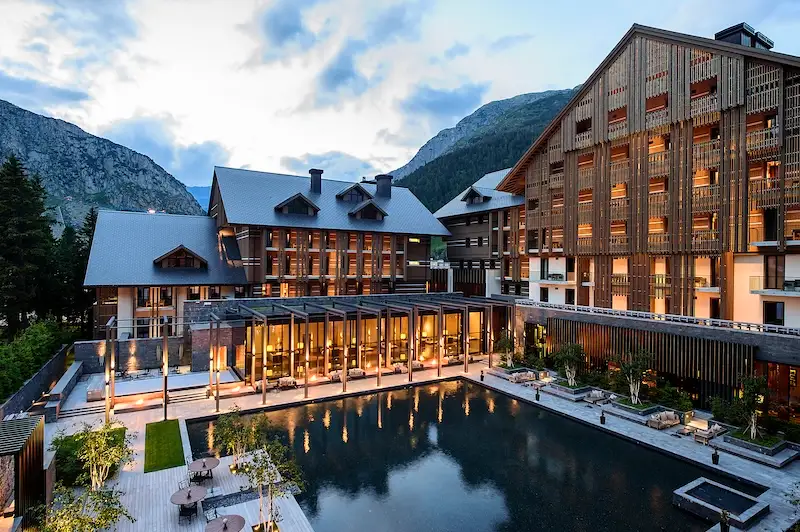 Hotel The Chedi Andermatt. Sicht in der Abenddämmerung auf den großen Outdoorpool und das erleuchtete Hotel mit Holzfassade. Im Hintergrund die Bergel