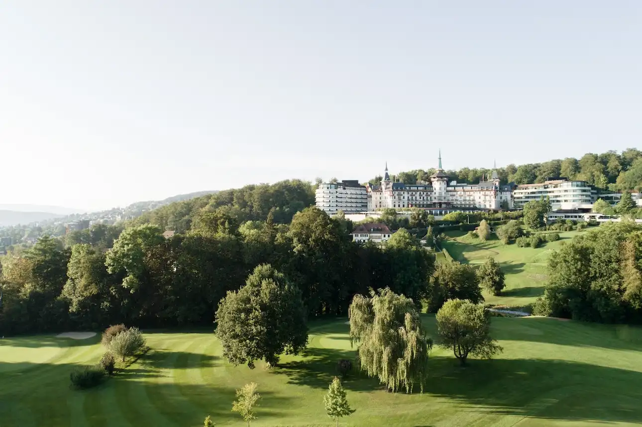 Hotel The Dolder Grand: Altes Hotel auf einem Hügel, davor grüne Wiesen und Wald mit blauem Himmel