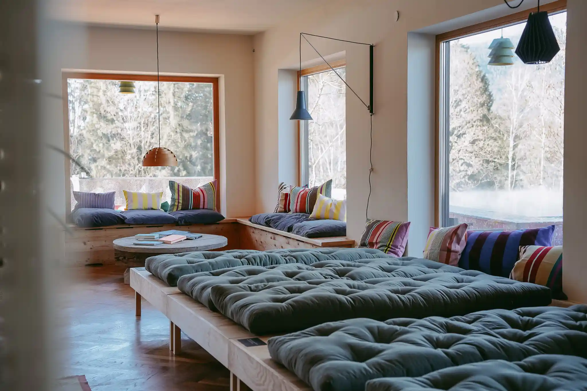 Der neue Badehosen-Ruheraum in Seebichl: Große Palettenkissen auf Holzliegen, bunte Kissen, große Fenster mit Blick auf Tannen