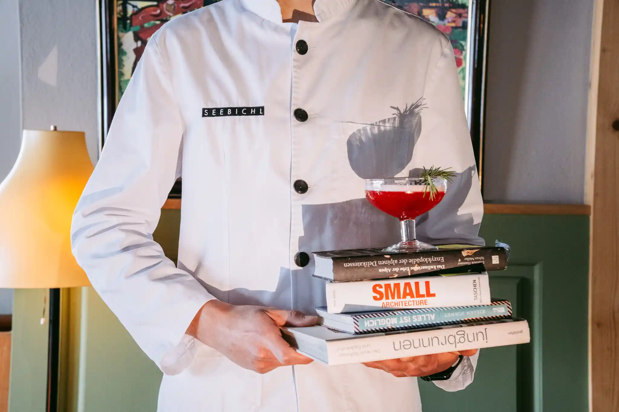 Books & Drinks: Mann mit weißer Serviceweste, der einen Stapel Bücher mit einem roten Cocktail darauf in den Händen hält