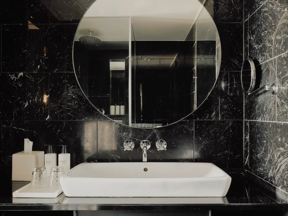 Badezimmer mit schwarzen Marmor-Vertäfelungen, rundem Spiegel und weißem Waschbecken im Hotel The Dean Munich