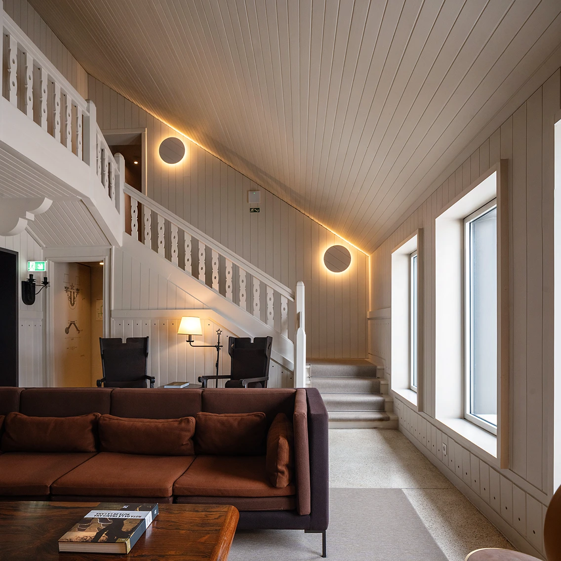 Gemütliche Kaminlounge mit rostbraunem Sofa, weiß gestrichener Holzvertäfelung an Decke und Wänden sowie einer eleganten Holztreppe.||©  Maria João Gala